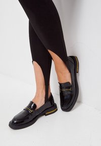 Mocassins en cuir verni noir avec un bout rond, des coutures d'accent jaune et une semelle plate. Portés avec des léggings noirs présentant des découpes.