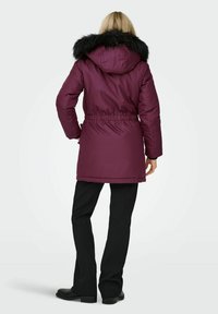 Parka isolata di un viola scuro con cappuccio in pelliccia sintetica, vita elastica e maniche lunghe. Abbinata a pantaloni neri e stivali alla caviglia.
