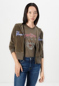 Maslinasto zelena velur zip-up dukserica sa plavim i crvenim izvezenim logotipima, u kombinaciji s smeškastim majicom s dizajnom tigra i plavim trapericama.