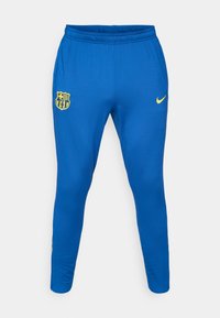 Μπλε αθλητικό παντελόνι από λείο ύφασμα, με κίτρινο λογότυπο της Nike στη δεξιά πλευ ρά και κίτρινο έμβλημα της FC Barcelona στην αριστερή πλευρά.
