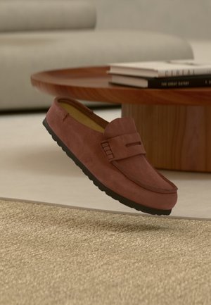 Mocassin en daim marron avec semelle noire flottant au-dessus d'un tapis beige dans un salon avec une table basse en bois et des livres empilés.