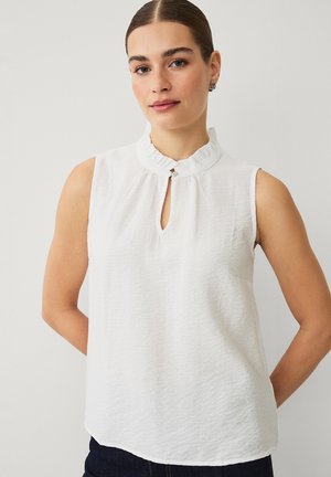 Kobieta z zaczesanymi do tyłu włosami, ubrana w białą, bezrękawnikową bluzkę z falbanianym kołnierzykiem i ozdobnym guzikiem w kształcie klucza, stojąca na jednolitym tle.