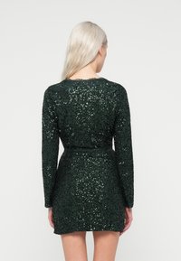 Πράσινο φόρεμα σε πούλια με μακριά μανίκια, στενή σιλουέτα και ζώνη στη μέση. Διαθέτει στρογγυλή λαιμόκοψη και υφή με γυαλιστερή επιφάνεια.