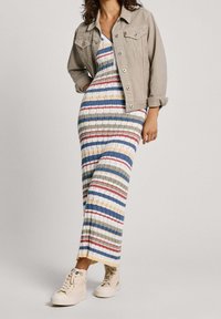 Maxiklänning med flerfärgade ränder i blått, grönt, rött och gult, kombinerad med en ljusbeige jeansjacka och krämfärgade sneakers.