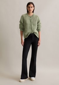 Hellgrüner gestrickter Pullover mit Lochmuster, Rundhalsausschnitt und gerippten Bündchen, kombiniert mit schwarzen Schlaghosen und weißen Plateauschuhen.