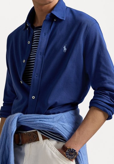 Blauwe overhemd met knoopsluiting en een textuurafwerking, met een geborduurd logo, gelaagd over een marineblauw en wit gestreept shirt. Gestyled met een lichtblauwe trui en een brown leren riem.