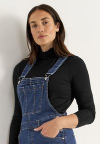 Jeansoveraller i medelblå färg med justerbara axelband, två framfickor och kontrasterande sömmar, ihop med en figursydd svart polotröja.