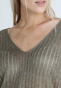 Pleťový pletený top s výstřihem do V z průsvitného olivově zeleného materiálu s vertikálními pruhy a texturovaným povrchem, který zdůrazňuje lehký design.