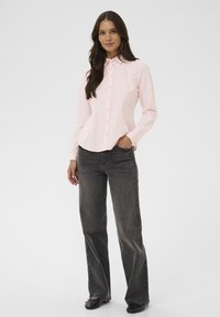 Chemise boutonnée rose clair avec col, manches longues et plis à l'avant, associée à un jean évasé gris foncé et des chaussures noires.