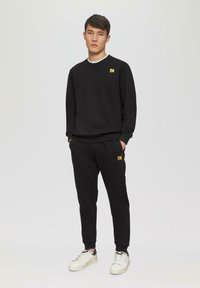 Sweat-shirt noir à col rond, avec un petit logo jaune sur la poitrine, associé à un pantalon de jogging noir et des baskets blanches.