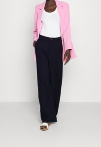 Jasnoróżowa marynarka na białym topie na ramiączkach, w zestawieniu z granatowymi szerokimi spodniami i białymi sandałami. Gładki materiał, luźny fason i brak widocznych wzorów.