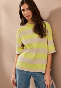 Prúžkovaný top s krátkym rukávom v svetlo béžovej a fluorescenčne žltej farbe, s voľným strihom a mäkkou látkou. Kombinovaný s modrými džínsami.