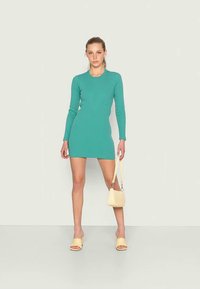 Vestido mini em turquesa com relevo, mangas longas e decote arredondado, combinado com chinelos amarelos e uma pequena bolsa. Textura lisa e ajustada.