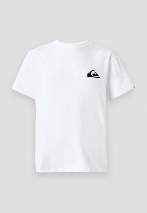 Junger Mann trägt ein weißes T-Shirt mit Logo, schwarze Hose, weiße Sneakers und einen schwarzen Bucket Hat und steht vor einem einfarbigen hellen Hintergrund.