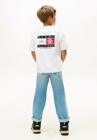 T-shirt bianca in cotone con loghi grafici neri e rossi sul retro, abbinata a jeans denim blu chiaro e scarpe sportive nere.