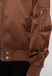 Bruine satijnen bomberjack met ribgebreide manchetten en zoom, zippered zak op de mouw, omrand logo detail en een gladde textuur.