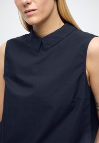 Camicia senza maniche blu navy con colletto a punta, realizzata in tessuto liscio, con tasca laterale e vestibilità rilassata.