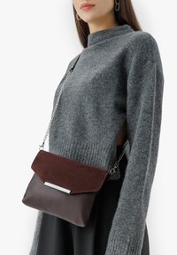 Borsa a tracolla in pelle marrone e suede bordeaux con accessori in metallo argentato e design a pattina piegata. Indossata con un maglione grigio.