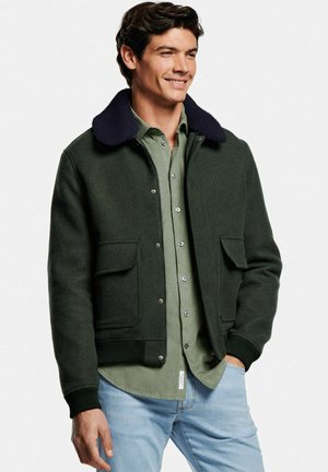 Grüne Wollmischjacke mit navyfarbenem Kragen, ausgestattet mit Druckknöpfen, gerippten Bündchen und Fronttaschen, getragen über einem hellgrünen Hemd.