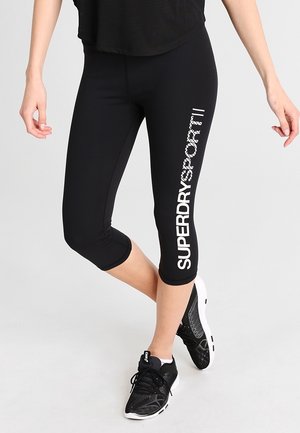 Kvinde iført sorte korte leggings med hvid tekst "SUPERDRY SPORT" på venstre ben og sorte træningssko med hvide såler.