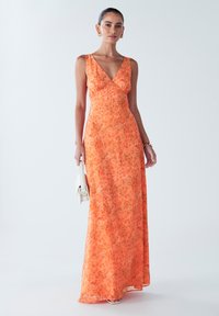 Dlouhé bezramínkové šaty v živé oranžové barvě s květinovým vzorem, s výstřihem ve tvaru V a splývavou látkou. Doplňují je malé bílé kabelky.