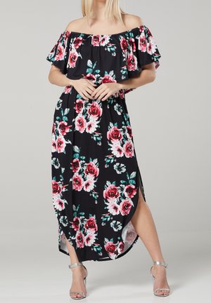 Kvinde iført sort off-shoulder kjole med pink og hvidt blomsterprint og sølvfarvede højhælede sandaler, stående med hænderne foldet.
