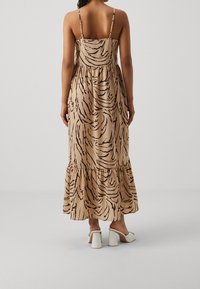 Vestido maxi en tela color tostado con un patrón abstracto negro, con un corpiño ajustado, dobladillo con volantes y tirantes delgados ajustables. Combinado con sandalias blancas de tacón cuadrado.