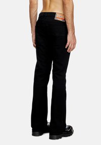 Pantalones de mezclilla negra con un diseño de pierna acampanada, que cuentan con dos bolsillos traseros y una etiqueta de marca naranja en la cinturilla. Textura suave.