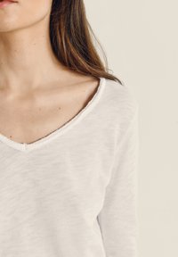 Žena s dlouhými hnědými vlasy, která má na sobě bílou dlouhý rukáv s výstřihem do V, ukazující svůj krk, rameno a horní část hrudníku na jednobarevném pozadí.