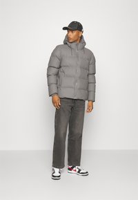 Giacca a vento grigia con zip frontale, cappuccio e dettagli elasticizzati; abbinata a jeans scuri e sneakers bianche e nere con accenti rossi.