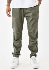 Pantalones jogger verde oliva con puños el�ásticos, bolsillos frontales, detalle de logo sutil y parches en las rodillas reforzados, combinados con zapatillas blancas.