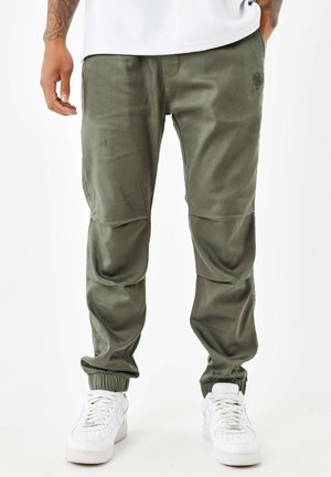 Pantalones jogger verde oliva con puños elásticos, bolsillos frontales, detalle de logo sutil y parches en las rodillas reforzados, combinados con zapatillas blancas.