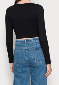 Schwarzes geripptes Langarm-Crop-Top, kombiniert mit blauen hochtaillierten Jeans, die über geschwungene Gesäßtaschen und ein „ROLLAS“-Label am Bund verfügen.