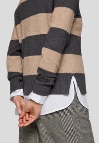 Gestreifter Pullover aus dunkelgrauer und beiger Wolle mit gerippten Bündchen und seitlichen Schlitzen. Kombiniert mit einem weißen Hemd mit Kragen und karierten Hosen.