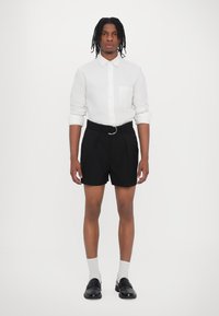 Fehér gombos ing, elülső zsebével, fekete elegáns shorttal és fekete loaferekkel párosítva. A modellen fehér bokazokni van.