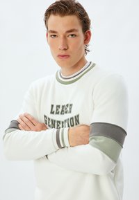 Vit sweatshirt med grå och grönrandiga muddar, med texten "LEADER GENERATION" i fet stil över bröstet.