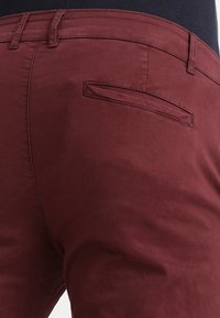 Primo piano di una persona che indossa pantaloni rosso scuro con tasca a filetto sul retro e passanti per cintura, abbinati a una maglietta nera.