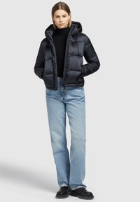 Giubbotto a vento nero con cappuccio, caratterizzato da un design trapuntato e chiusura con zip. Indossato sopra un dolcevita nero e abbinato a jeans azzurri chiari.