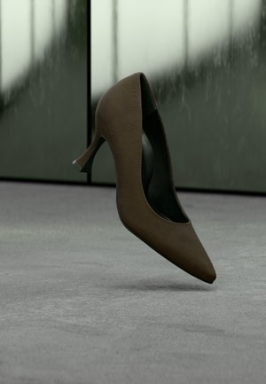 Une seule chaussure à talon haut marron foncé suspendue au-dessus d'un sol gris avec des panneaux de verre flous en arrière-plan.