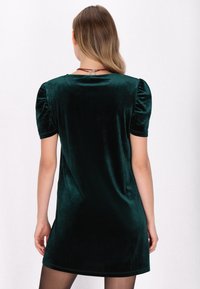 Grünes Samtkleid mit kurzen Puffärmeln, knielang, glatte Textur und ein einfaches Design, getragen über transparenten schwarzen Strumpfhosen.