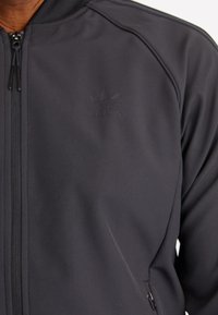 Černá zipová bunda vyrobená z hladké tkaniny, s tónovým logem adidas na hrudi a jemnými detaily švů. Světlejší akcenty u zipů.