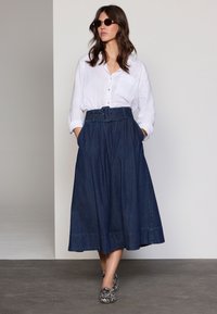 Denim rok met plooien, hoge taille en een brede riem. Witte blouse met knopen en gestreepte slip-on schoenen. Accessoires omvatten een zonnebril.