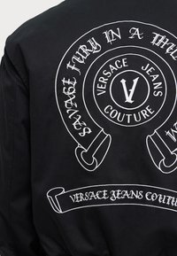 Melns žakets ar baltu izšūtu grafiku uz muguras, kurā ir teksts "Versace Jeans Couture" un apļveida dizaina elementi.