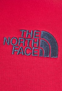 Czerwony materiał o gładkiej fakturze z granatowym haftowanym logo "THE NORTH FACE" oraz wzorem półkopuły.