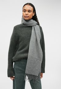 Harmaa villahuivi, jossa on liehukkeet reunoissa, esittelee vaaleasta tummempaan gradienttiin, päällä tummanvihreä neulepusero ja farkut, yksinkertaista taustaa vasten.