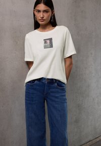 Camiseta color crema con un gráfico cuadrado en la parte delantera, combinada con jeans de pierna ancha de color azul oscuro. La textura parece suave y casual.