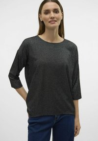 Top nero a coste con maniche a tre quarti, scollatura rotonda e leggero riflesso. Abbinato a jeans in denim blu, su uno sfondo neutro.