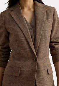 Ruskea herringbone-blazeri, jossa on aukkopeitteiset läpät, kaksi etutaskua ja kolmonen napitus. Materiaali näyttää olevan teksturoitu ja istuvuus on räätälöity.
