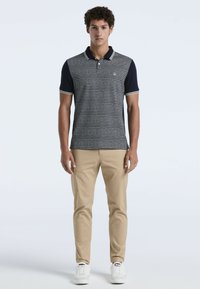 Polo shirt à motifs bleu marine et blanc avec un col, des manches courtes et des poignets rayés, associé à un pantalon beige et des baskets blanches.