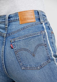Jean en denim bleu clair avec une étiquette de marque en cuir, présentant un design distinctif de poche arrière avec des surpiqûres orange et une étiquette logo rouge.